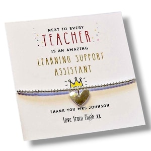 May include: A silver beaded bracelet with a small heart charm attached to a white display card. The card features the text: NEXT TO EVERY TEACHER IS AN AMAZING LEARNING SUPPORT ASSISTANT, a yellow crown icon, THANK YOU MRS JOHNSON, and Love from Elijah xx.