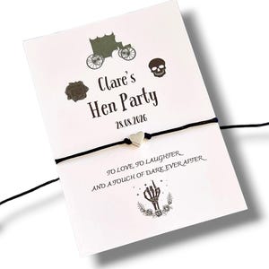 Puede incluir: Una tarjeta blanca con el texto "Clare's Hen Party 28.08.2026" e ilustraciones de un carruaje, una calavera y una rosa. Una pulsera de cordón negro con un dije de corazón plateado está adjunta.