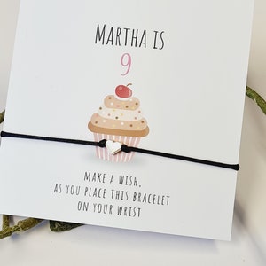 May include: A birthday-themed card with the text "MARTHA IS 9" and a cupcake illustration. A black string bracelet with a small heart charm is in front of the card. The card also reads "MAKE A WISH, AS YOU PLACE THIS BRACELET ON YOUR WRIST."