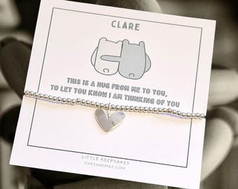 May include: A silver bracelet with a heart charm and a card that reads "CLARE THIS IS A HUG FROM ME TO YOU. TO LET YOU KNOW I AM THINKING OF YOU LITTLE KEEPSAKES CHEZANDMAZ.COM". The card features a cartoon illustration of two animals hugging.