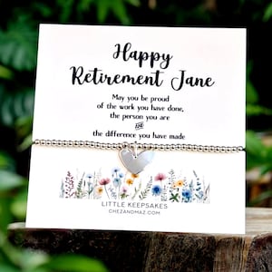 May include: A silver beaded bracelet with a central heart charm displayed on a white card featuring the text Happy Retirement Jane, a sentimental message, and a colorful floral illustration at the bottom with the brand name Little Keepsakes.