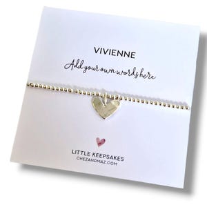 Könnte beinhalten: Ein silbernes Perlenarmband mit Herzanhänger, präsentiert auf einer weißen Karte. Die Karte trägt den Namen "VIVIENNE" und den Text "Add your own words here". Der Markenname "LITTLE KEEPSAKES" und die Website-Adresse sind ebenfalls sichtbar.