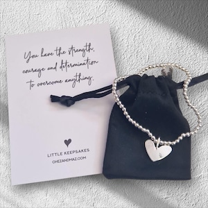 May include: A silver beaded bracelet with a heart charm. The bracelet comes in a black velvet pouch with a card that reads "You have the strength, courage and determination to overcome anything." and "LITTLE KEEPSAKES CHEZANDMAZ.COM"