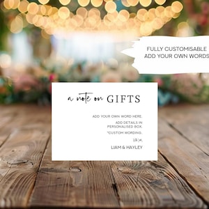 May include: A white card with black text that reads "a note on GIFTS". The text below reads "ADD YOUR OWN WORD HERE. ADD DETAILS IN PERSONALISED BOX. 'CUSTOM WORDING.' LIAM & HAYLEY". The card is on a wooden table with a blurred background of lights.