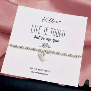 May include: A silver beaded bracelet with a heart charm hangs in front of a white card. The card reads "LIFE IS TOUGH but so are you" in black text. The card also has the name "Kellie" and the website "chezandmaz.com".