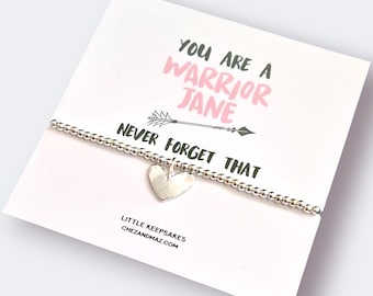 May include: A silver beaded bracelet with a heart charm is displayed on a white card. The card features the text "YOU ARE A WARRIOR JANE NEVER FORGET THAT" in pink and black. The brand name "LITTLE KEEPSAKES" is also visible.