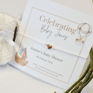 Op de afbeelding: Een babyborrelkaart met de tekst "Celebrating Baby Jones" en "Joanne's Baby Shower 4th February 2024". Een witte armband met een hartvormige bedel ligt op de kaart. Een klein gebreid wit babyschoentje is zichtbaar.