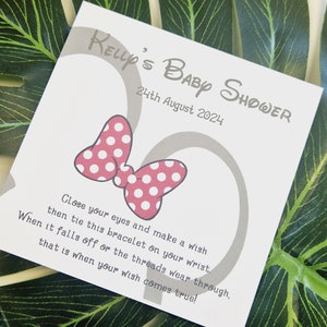 May include: A white card with gray text and a pink bow with white polka dots. The text reads "Kelly's Baby Shower 24th August 2024" and "Close your eyes and make a wish then tie this bracelet on your wrist. When it falls off or the threads wear through, that is when your wish comes true!"