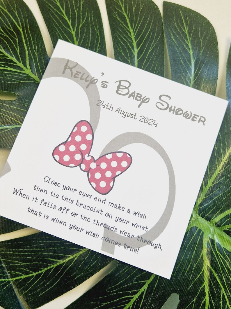 May include: A white card with gray text and a pink bow with white polka dots. The text reads "Kelly's Baby Shower 24th August 2024" and "Close your eyes and make a wish then tie this bracelet on your wrist. When it falls off or the threads wear through, that is when your wish comes true!"