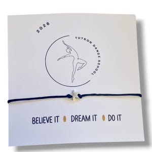 Peut inclure: Un bracelet en cordon bleu marine avec un charm en forme d'étoile argentée est présenté sur une carte blanche. La carte affiche le texte "2026" et le logo de l'école de danse Tutron. Les mots "BELIEVE IT DREAM IT DO IT" sont imprimés en dessous.