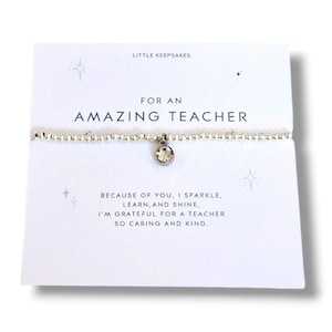 May include: A silver-toned beaded bracelet with a single clear round crystal charm, presented on a white card labelled LITTLE KEEPSAKES FOR AN AMAZING TEACHER, featuring the text: BECAUSE OF YOU, I SPARKLE, LEARN, AND SHINE, I'M GRATEFUL FOR A TEACHER SO CARING AND KIND.