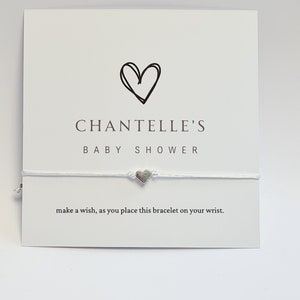Może przedstawiać: Biała bransoletka ze sznurka z zawieszką w kształcie srebrnego serca. Bransoletka jest dostarczana z kartką, na której napisano "CHANTELLE'S BABY SHOWER" i "zrób życzenie, zakładając tę bransoletkę na nadgarstek."
