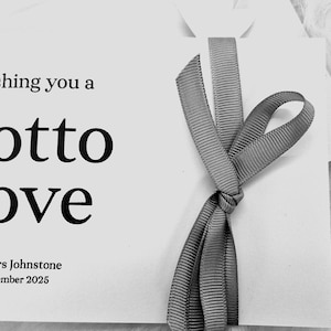 Könnte beinhalten: Eine weiße Karte mit dem Text "wishing you a lotto love" in Schwarz, gebunden mit einem grauen Band. Die Karte enthält auch die Namen "Mr & Mrs Johnstone" und das Datum "15th December 2026".