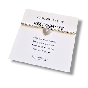 May include: A silver-coloured beaded bracelet with a heart-shaped charm attached to a white display card. The card features the text: CLARE, HERE'S TO THE NEXT CHAPTER. Behind you, all your memories. Before you, all your dreams. Around you, all who love you. Within you, all you need. LITTLE KEEPSAKES CHEZANDMAZ.COM.