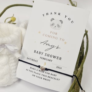 May include: A baby shower invitation card with a panda illustration, the text "Thank You For Coming To Amy's Baby Shower February 3, 2024". A black bracelet with a heart charm is attached. A small white bag is in the background.