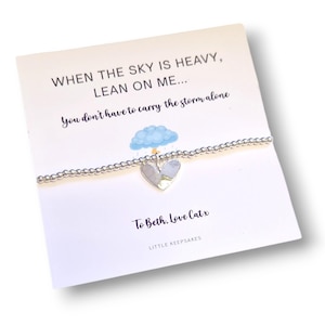 Op de afbeelding: Een zilveren armband met een hartvormige hanger, gepresenteerd op een witte kaart. De kaart bevat de tekst: "WHEN THE SKY IS HEAVY, LEAN ON ME... You don't have to carry the storm alone." Er is ook een kleine wolkenillustratie.