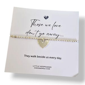 May include: A silver-toned beaded bracelet with a central heart-shaped charm, displayed on a white card featuring the text "Those we love don't go away... They walk beside us every day." and "LITTLE KEEPSAKES CHEZANDMAZ.COM" with a small heart icon.