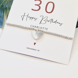 Op de afbeelding: Een witte kaart met de tekst "30 Happy Birthday Charlotte" in rood en zwart. Een zilveren armband met een hartbedel ligt op de kaart. De kaart heeft ook een klein rood hart en de tekst "Chezandmaz UK" in zwarte letters.