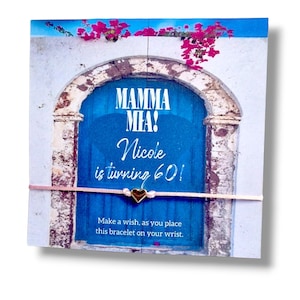 Pode incluir: Uma pulseira de fio rosa claro fino com um pequeno pingente de coração dourado presa a um cartão quadrado. O cartão apresenta uma porta azul em arco, flores rosas e o texto: Mamma Mia! Nicole is turning 60! Make a wish, as you place this bracelet on your wrist.