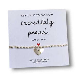 Könnte beinhalten: Ein silbernes Perlenarmband mit einem herzförmigen Anhänger auf einer weißen Karte. Die Karte trägt den Text: ABBY, JUST TO SAY HOW incredibly proud I AM OF YOU, zusammen mit einem kleinen roten Herzsymbol und LITTLE KEEPSAKES CHEZANDMAZ.COM.
