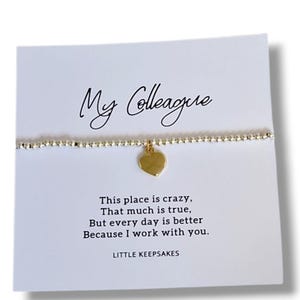 Op de afbeelding: Een fijne armband van zilverkleurige kraaltjes met een kleine gouden hartvormige bedel, gepresenteerd op een witte kaart met de tekst My Colleague, This place is crazy, That much is true, But every day is better Because I work with you, LITTLE KEEPSAKES.