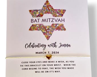 May include: A white card with a Star of David design filled with colorful flowers, the words "BAT MITZVAH" and "Celebrating with Jenna" are printed on the card. A gold heart charm is attached to a white cord.