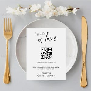 May include: A table setting featuring a white plate, gold cutlery, and a card with a QR code. The card says "Capture the love" and invites guests to share photos. A floral arrangement is in the background.