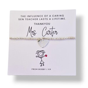 Puede incluir: Una pulsera de cuentas plateadas con un dije de corazón se muestra en una tarjeta blanca. La tarjeta tiene el texto: "THE INFLUENCE OF A CARING SEN TEACHER LASTS A LIFETIME. THANKYOU Mrs Carter FROM BOBBY J XX". También hay un dibujo de un muñeco de palitos con un corazón.