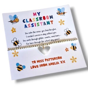 May include: A white card with the text "MY CLASSROOM ASSISTANT" in colourful letters, decorated with cartoon bees and flowers. A silver beaded bracelet with a heart charm is attached. The card reads "TO MISS PATTERSON LOVE FROM AMELIA XX."