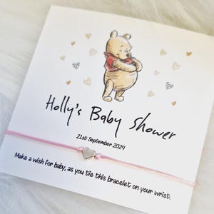 Peut inclure: Un bracelet de fête prénatale rose et blanc avec un charme en forme de cœur argenté. La carte indique "Holly's Baby Shower 21st September 2024" et "Faites un vœu pour le bébé, en attachant ce bracelet à votre poignet."