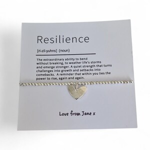 May include: A silver bracelet with a heart charm hangs below a white card. The card reads "Resilience" with a definition and the phrase "Love from Jane x". The bracelet is on a white background.