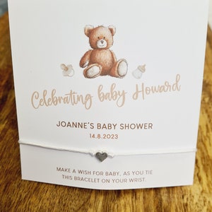 May include: White string bracelet with a silver heart charm. The card attached to the bracelet reads "Celebrating baby Howard, Joanne's Baby Shower, 14.8.2023. Make a wish for baby, as you tie this bracelet on your wrist."