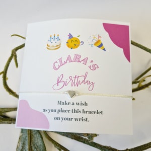Op de afbeelding: Een witte kaart met roze accenten en een zilveren armband met een hartbedel. De kaart heeft een verjaardagstaart, een feestmuts en een feesttoeter illustraties. De tekst op de kaart luidt "CLARA'S Birthday" en "Maak een wens terwijl je deze armband om je pols doet."