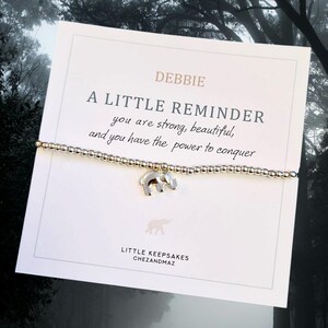 May include: A silver bracelet with a small elephant charm and a card that reads "A Little Reminder you are strong, beautiful, and you have the power to conquer". The card is signed "Debbie" and has the text "Little Keepsakes Chezandmaz" at the bottom.