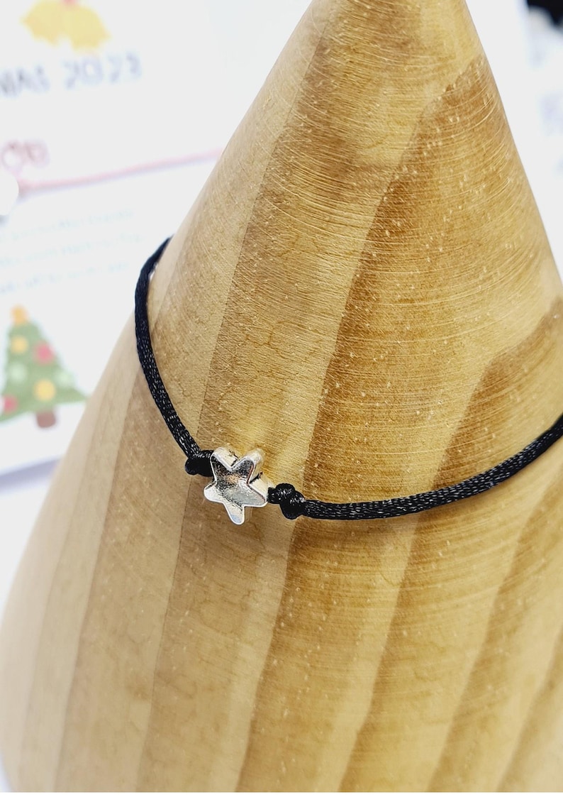May include: A black cord bracelet with a small, silver-coloured star charm. The bracelet is knotted on either side of the star. The bracelet is displayed on a wooden cone-shaped object.