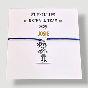 May include: A white card featuring the text "ST PHILLIPS NETBALL TEAM 2025 JOSIE" and a netball player stick figure. A blue cord bracelet with a silver heart charm is attached to the card.
