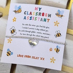 Può includere: Un braccialetto argentato con perline e un ciondolo a forma di cuore è esposto su una carta bianca. La carta recita "MY CLASSROOM ASSISTANT" con una poesia e "LOVE FROM ISLAY XX". La carta è decorata con illustrazioni di api e fiori.