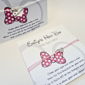 May include: A pink and white polka dot bow with a silver charm that says "Bride to Be" on a white string bracelet. The bracelet is tied in a bow and has a card attached to it that says "Emily's Hen Do 29th April 2023".