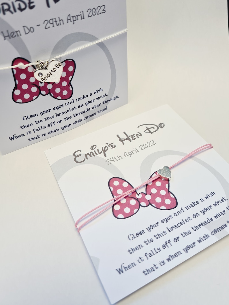 May include: A pink and white polka dot bow with a silver charm that says "Bride to Be" on a white string bracelet. The bracelet is tied in a bow and has a card attached to it that says "Emily's Hen Do 29th April 2023".