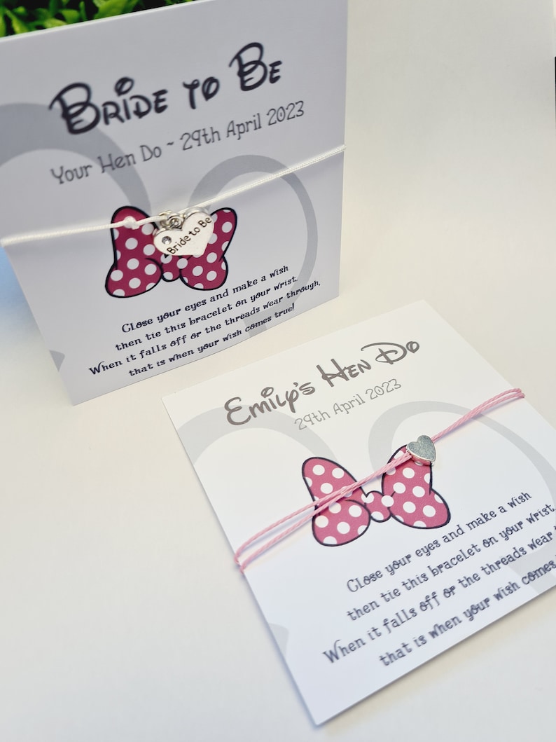May include: Two white cards with pink and white polka dot bows and silver heart charms. The cards have text that reads "Bride to Be Your Hen Do - 29th April 2023" and "Emily's Hen Do 29th April 2023". The text on the cards instructs the recipient to make a wish and tie the bracelet on their wrist. When the bracelet falls off or the threads wear through, their wish will come true.