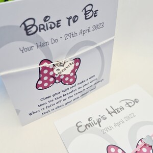 May include: A white card with the text "Bride to Be" and "Your Hen Do - 29th April 2023". A pink and white polka dot bow with a silver charm that says "Bride to Be" is attached to the card with a white string. The card also includes instructions for a wish bracelet.