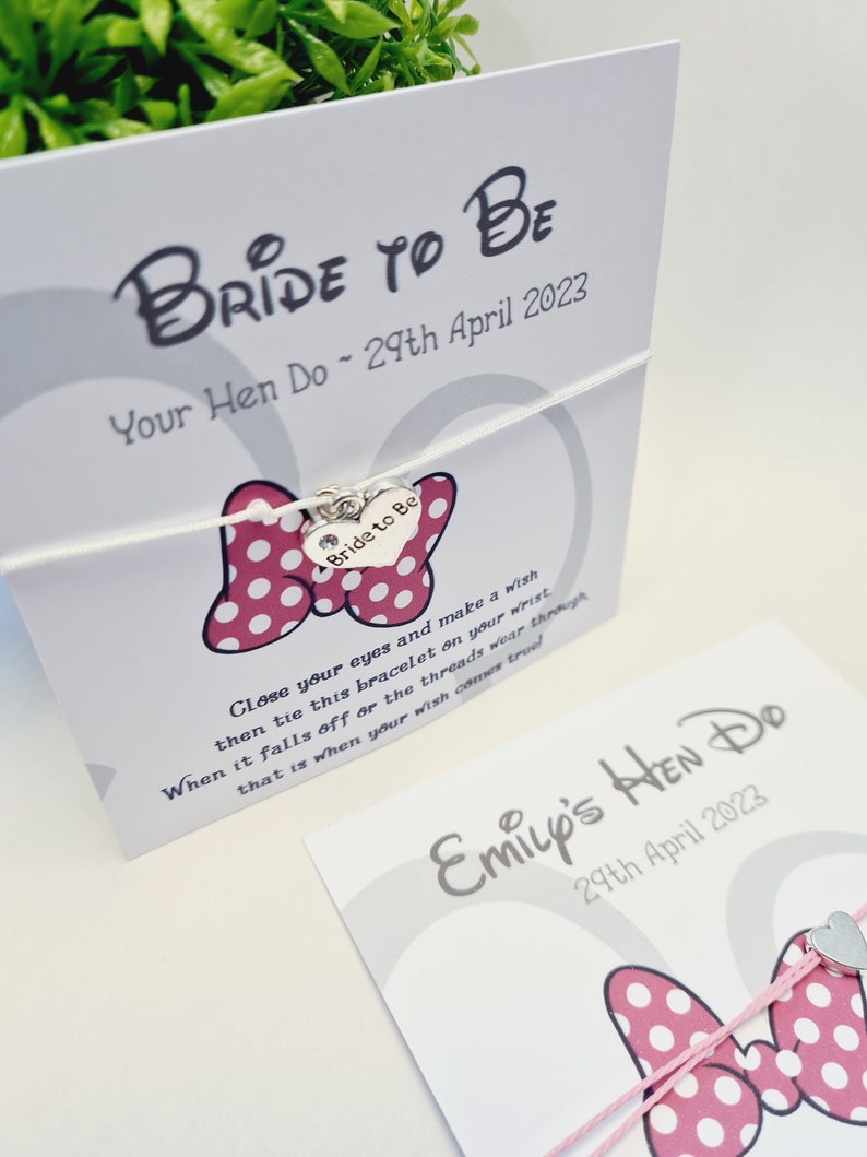 May include: A white card with the text "Bride to Be" and "Your Hen Do - 29th April 2023". A pink and white polka dot bow with a silver charm that says "Bride to Be" is attached to the card with a white string. The card also includes instructions for a wish bracelet.