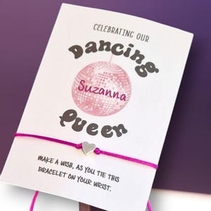 Puede incluir: Una bola de discoteca rosa y plateada con el nombre "Suzanna" escrito en ella. El texto "Celebrating our Dancing Queen" está por encima de la bola de discoteca y el texto "Make a wish, as you tie this bracelet on your wrist." está por debajo de la bola de discoteca. Un brazalete de cuerda rosa con un dije de corazón plateado está unido a la tarjeta.