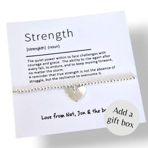May include: A silver beaded bracelet with a central heart charm displayed on a white card. The card features the definition of Strength and the text Love from Nat, Jon & the br, along with a circular graphic that reads Add a gift box.