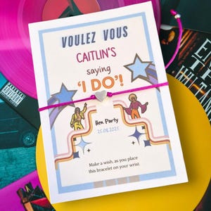 Puede incluir: Una tarjeta blanca con el texto "VOULEZ VOUS CAITLIN'S saying 'I DO'!" y "Hen Party 25.08.2025". Una pulsera rosa con un dije de corazón plateado adjunta. La tarjeta tiene un diseño retro con ilustraciones.