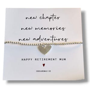 Op de afbeelding: Een zilveren armband met een hartvormige bedel, gepresenteerd op een witte kaart. De kaart bevat de tekst "new chapter, new memories, new adventures" en "HAPPY RETIREMENT MUM". Een klein rood hartje is ook zichtbaar.