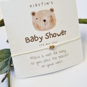 May include: A white card with brown text that reads "KIRSTIN'S Baby Shower 7TH MAY 2024" and a cartoon bear illustration. A white string bracelet with a gold heart charm is attached to the card. The text on the card reads "make a wish for baby, as you place this bracelet on your wrist."