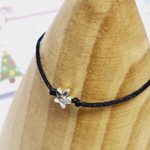 May include: A black cord bracelet with a small, silver-coloured star charm. The bracelet is knotted on either side of the star. The bracelet is displayed on a wooden cone-shaped object.