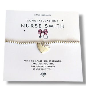 Pode incluir: Uma pulseira de contas prateadas com um pingente de coração num cartão branco. O cartão apresenta o texto LITTLE KEEPSAKES, CONGRATULATIONS NURSE SMITH, uma ilustração de um laço rosa e um estetoscópio, e uma mensagem sobre compaixão e força.