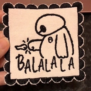 Pode incluir: Um remendo de tecido branco com bordado preto de um robô de desenho animado segurando uma chave e a palavra "BALALALA" embaixo.